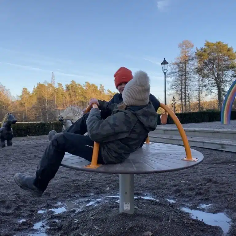 Två barn leker på en snurrkarusell på en lekplats. Det är vinter och himlen är blå.